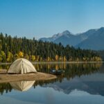 Tenda da campeggio bianca, accanto ad un lago con dietro un bosco fitto e una montagna