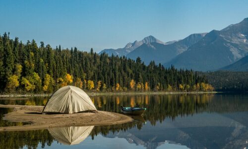 Tenda da campeggio bianca, accanto ad un lago con dietro un bosco fitto e una montagna
