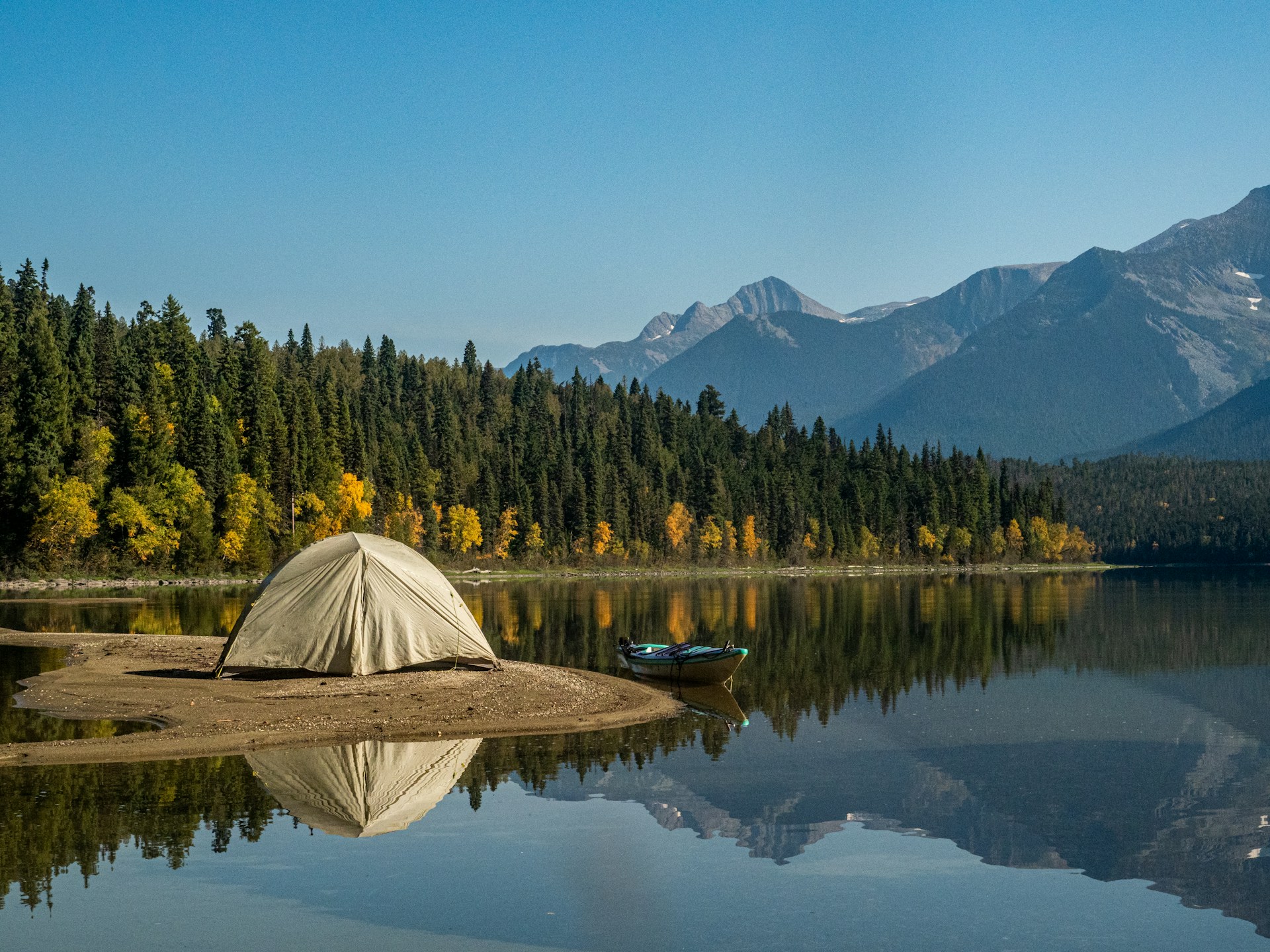 Tenda da campeggio bianca, accanto ad un lago con dietro un bosco fitto e una montagna