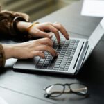 Mani di una donna che digitano sulla tastiera di un computer portatile grigio posizionato su una scrivania scura con un paio di occhiali da vista in primo piano