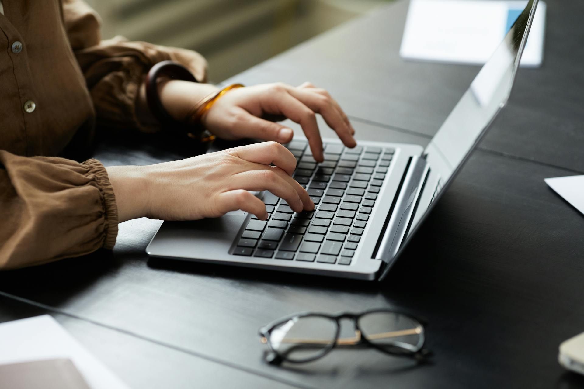 Mani di una donna che digitano sulla tastiera di un computer portatile grigio posizionato su una scrivania scura con un paio di occhiali da vista in primo piano