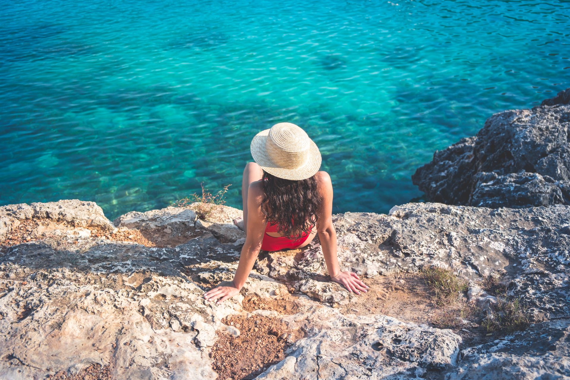 Donna con cappello di paglia seduta sugli scogli che guarda il mare cristallino di Bisceglie in Puglia.