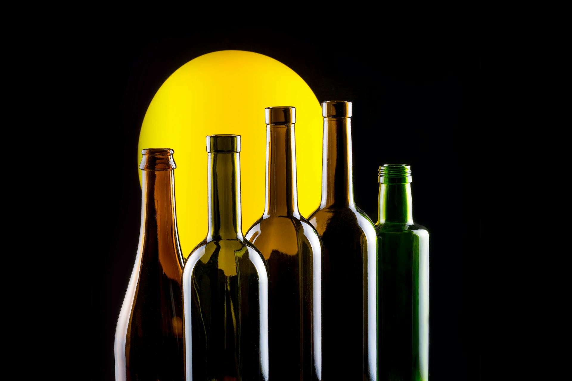 Foto still life di bottiglie di vetro per vino e olio su sfondo scuro con luce gialla circolare.